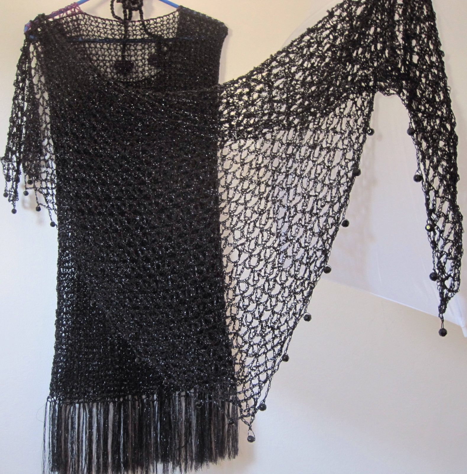 Black lurex crochet shawl draped over a black fringed dress on a hanger, side view of the shimmering wrap.