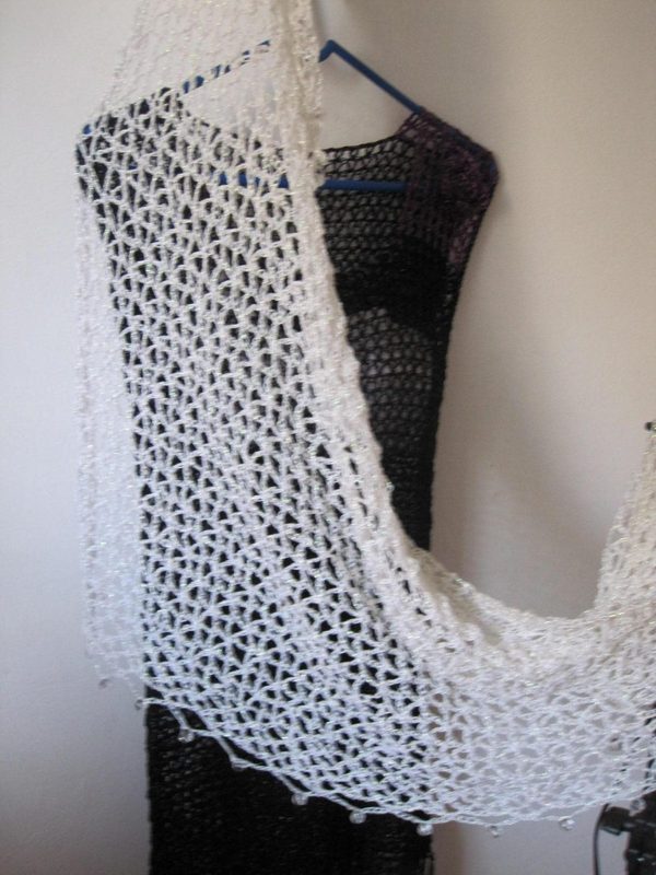 Cream crochet shawl draped over a black dress on a hanger, side view of the airy openwork wrap.