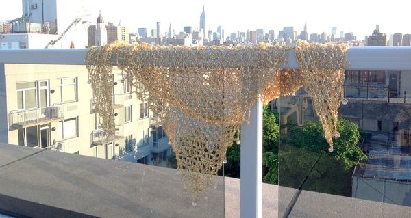 Gold lurex crochet shawl draped over a glass rooftop railing, with the New York City skyline stretching behind it in daylight.