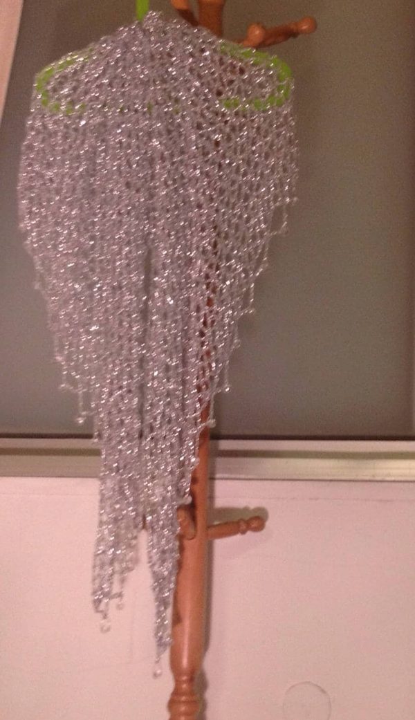 Silver beaded lurex crochet shawl hanging full-length from a wooden coat stand, its shimmering openwork weave and beaded edge catching the light.