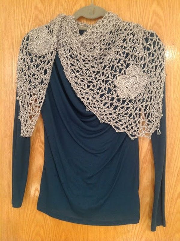 Silver lurex shawl with crocheted flower appliques draped over a navy blue top on a hanger, wooden door behind.